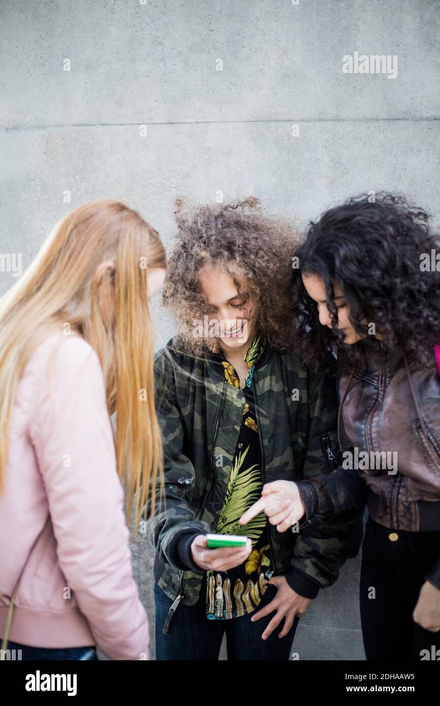 Smiling teenage boy showing smart phone to female friends against wall Stock Photo