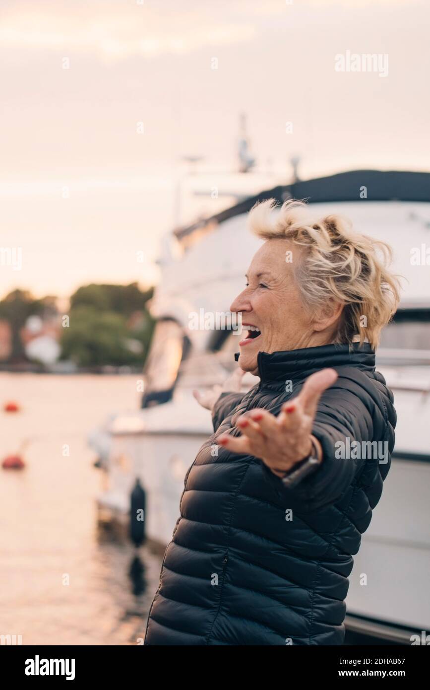 Side view of excited senior woman standing with arms outstretched at harbor Stock Photo