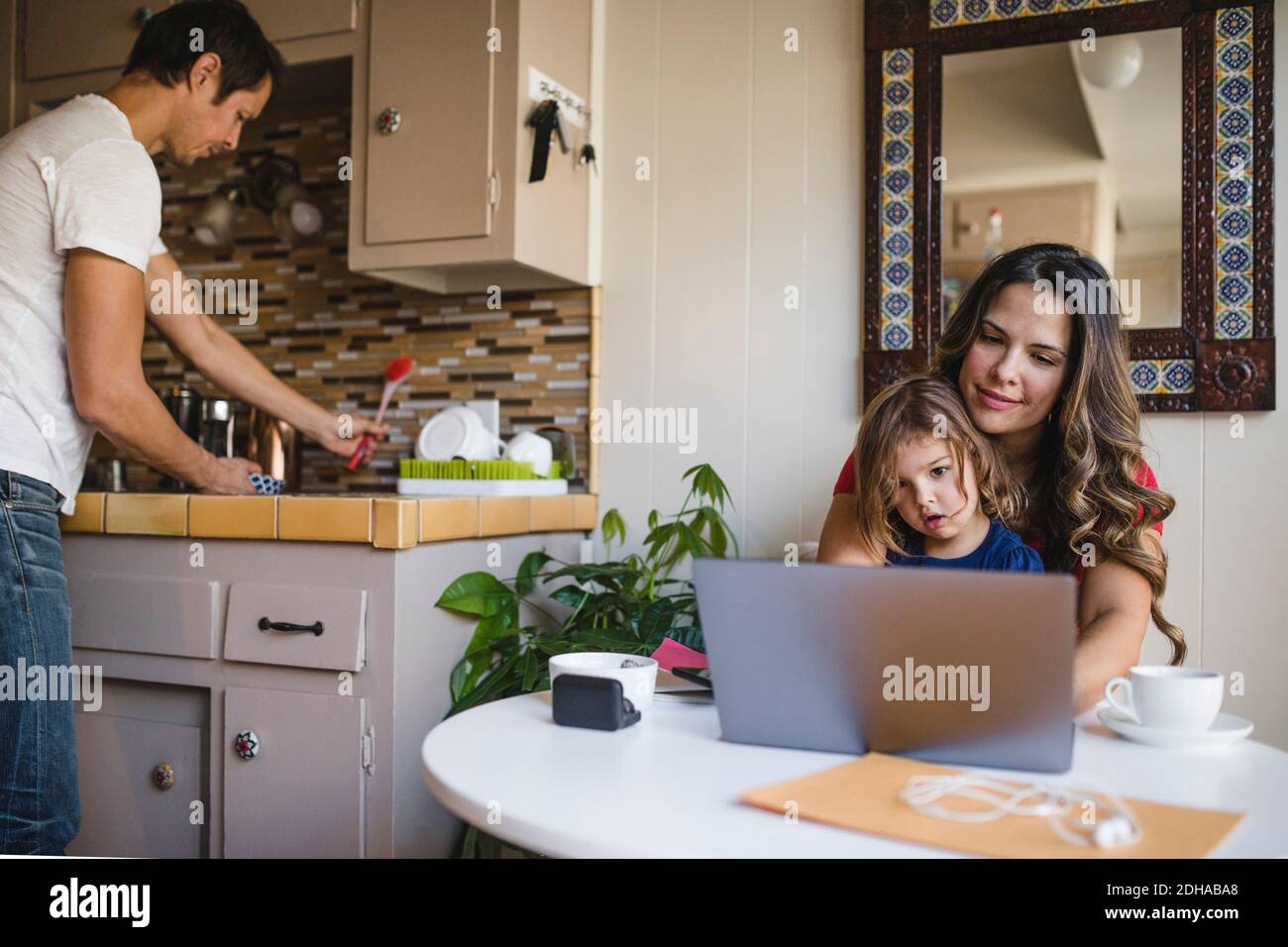 Mother and daughter using laptop while father working in kitchen Stock Photo