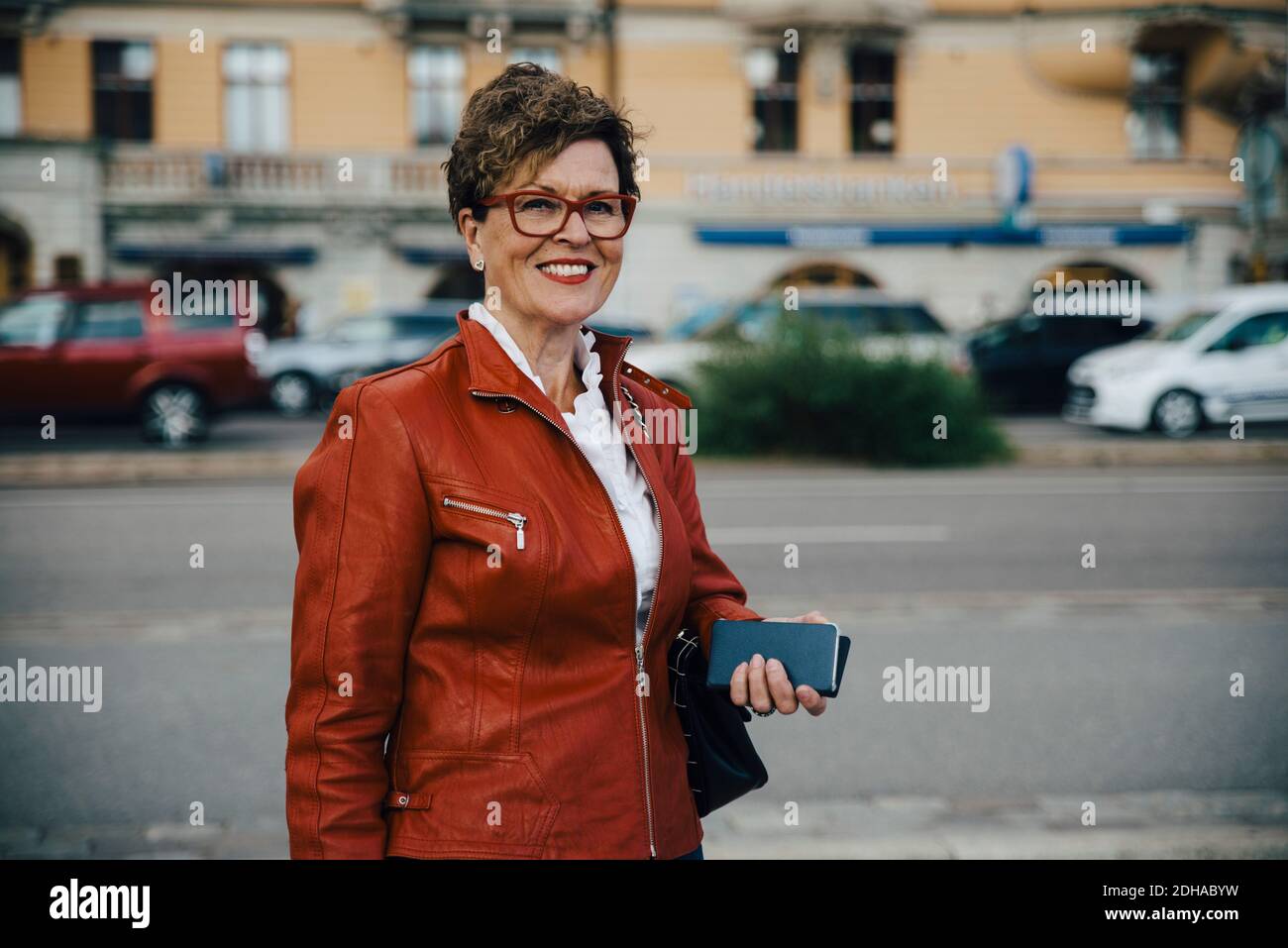 Portrait of smiling senior woman wearing brown jacket while standing by city street Stock Photo