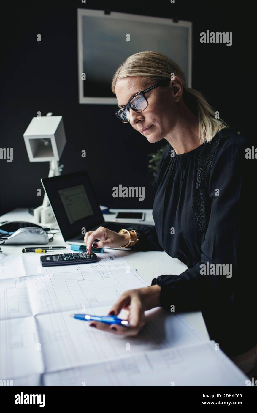 Businesswoman looking at blueprint while using calculator on desk Stock Photo
