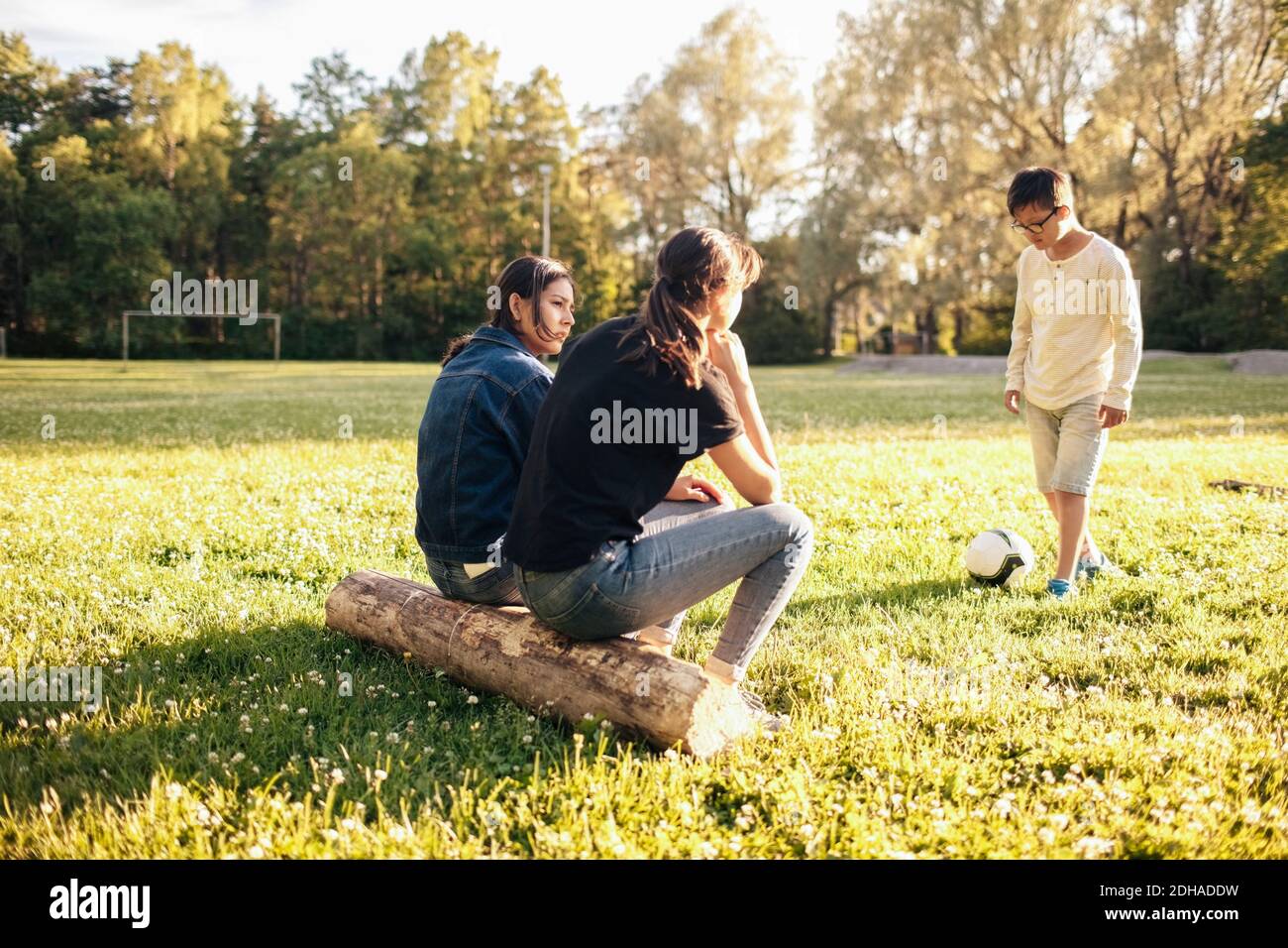 Sisters sitting on log while boy playing with ball at park during sunny day Stock Photo