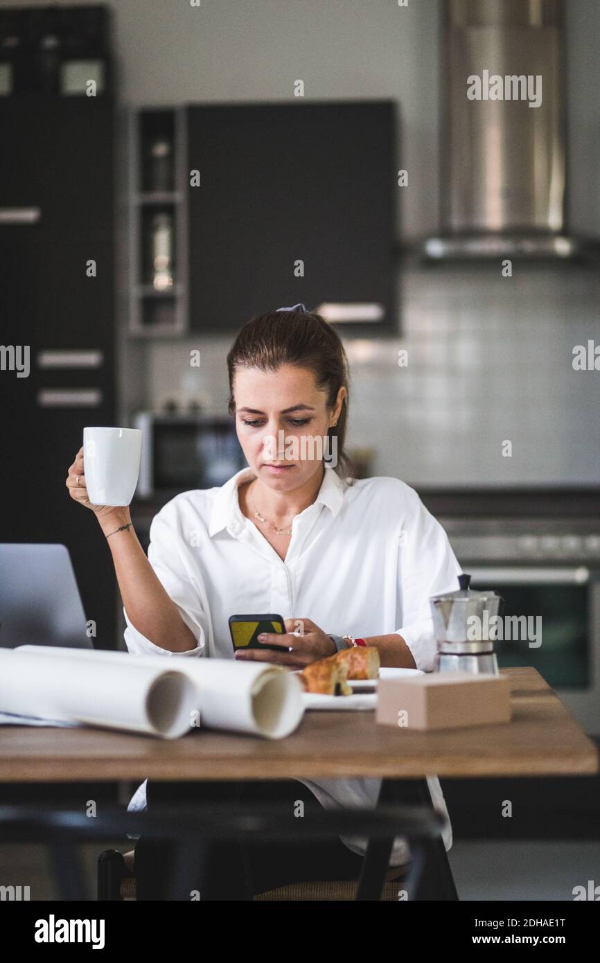 Female freelancer using phone while sitting with coffee cup at home Stock Photo