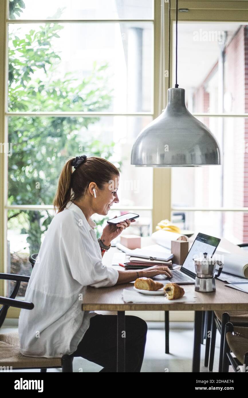 Smiling female freelancer using laptop while talking through smart phone at home office Stock Photo