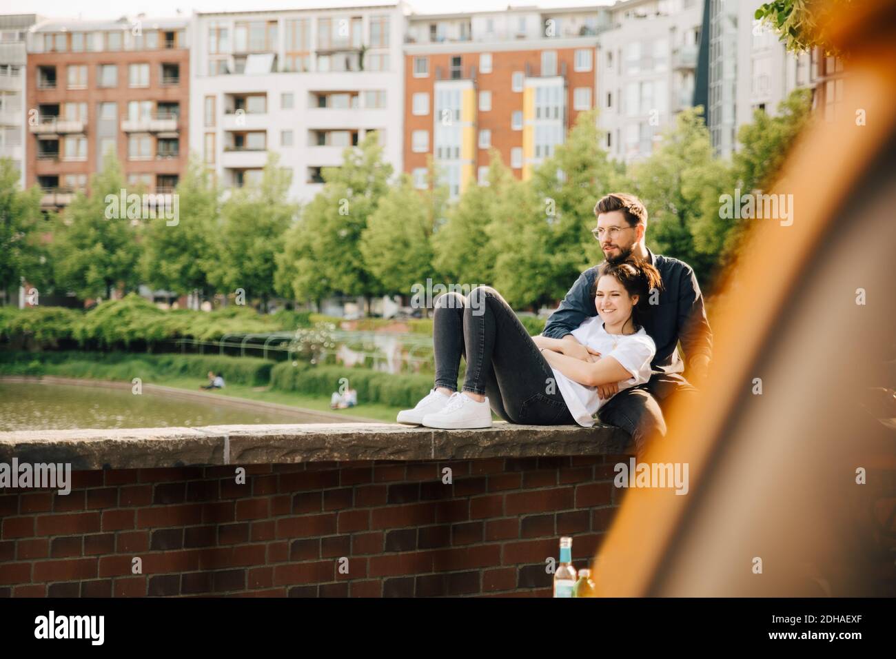 Young man with smiling female partner looking away while sitting on retaining wall Stock Photo