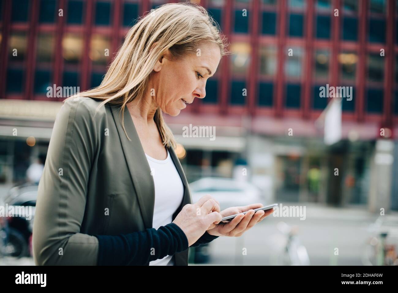 Blond mature businesswoman using smart phone against building in city Stock Photo
