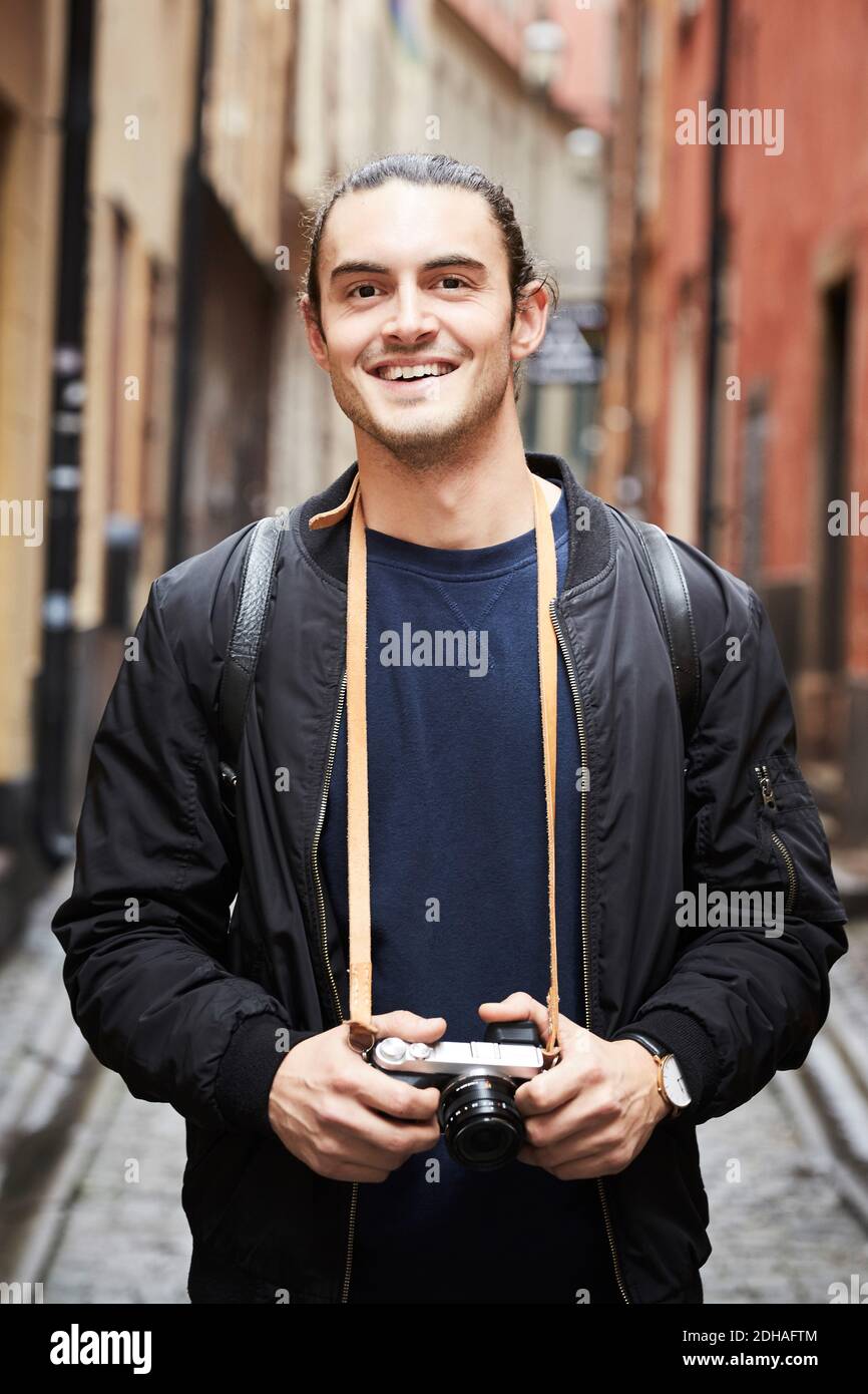 Portrait of smiling young man holding camera while standing in alley Stock Photo