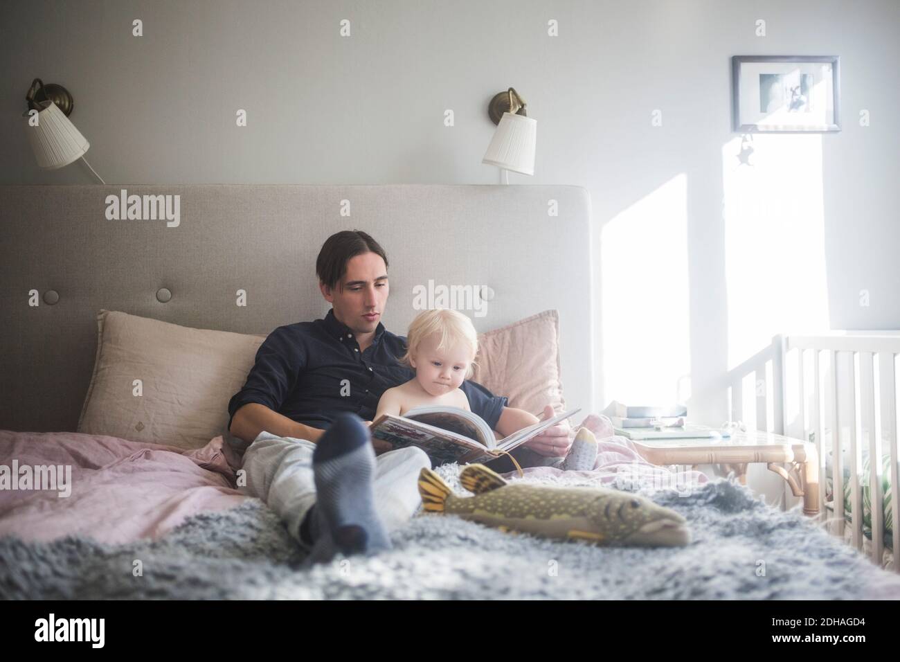 Father reading picture book with toddler son while sitting on bed at home Stock Photo
