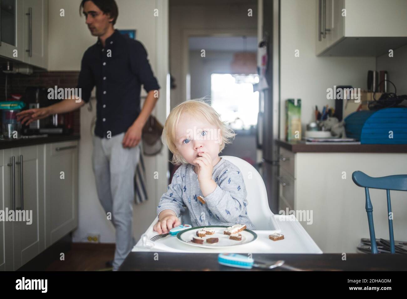 Portrait of blond baby boy eating while sitting on high chair at kitchen with father in background Stock Photo