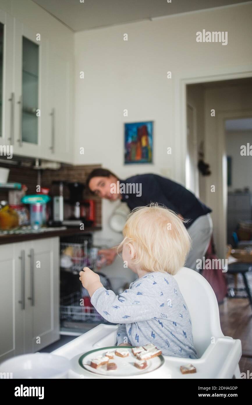 Blond baby boy sitting on high chair while looking at father in kitchen Stock Photo