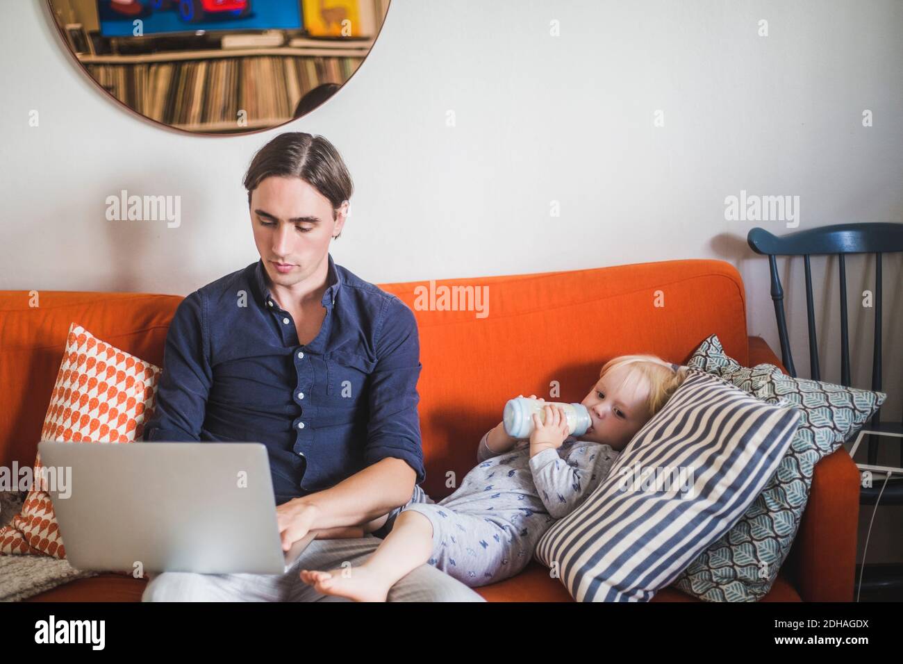 Stay at home father using laptop while sitting by toddler son drinking milk from bottle on sofa in apartment Stock Photo