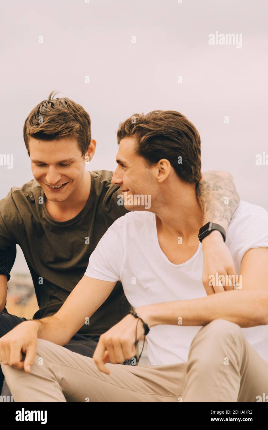 Smiling male friends sitting with arm around against sky Stock Photo