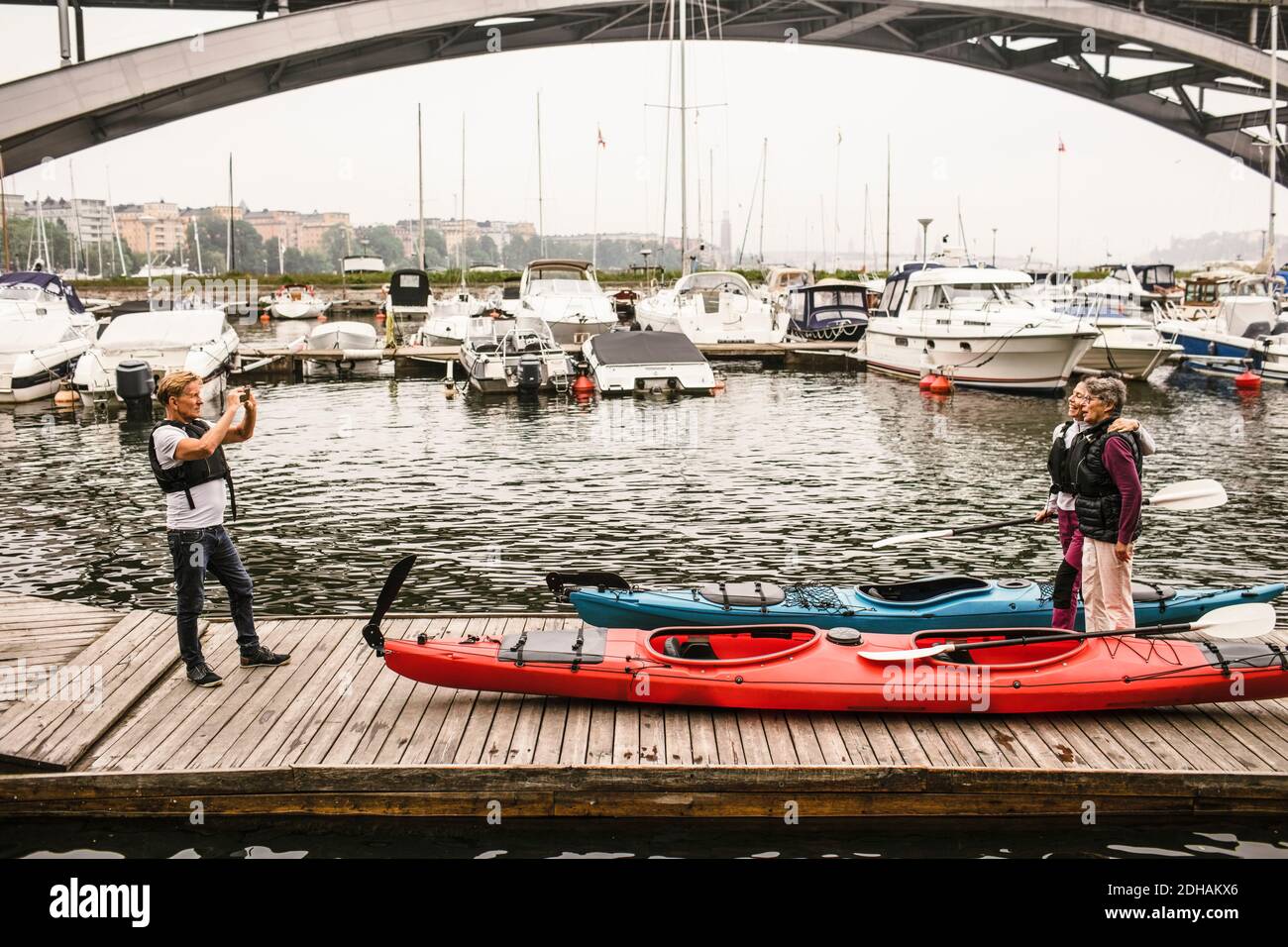 Senior male photographing female friends on jetty during kayaking course Stock Photo