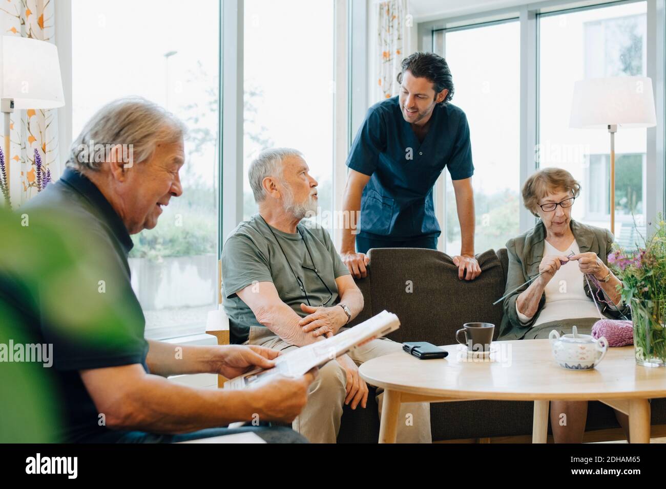 Male healthcare worker talking to senior man sitting on sofa amidst friends at retirement home Stock Photo