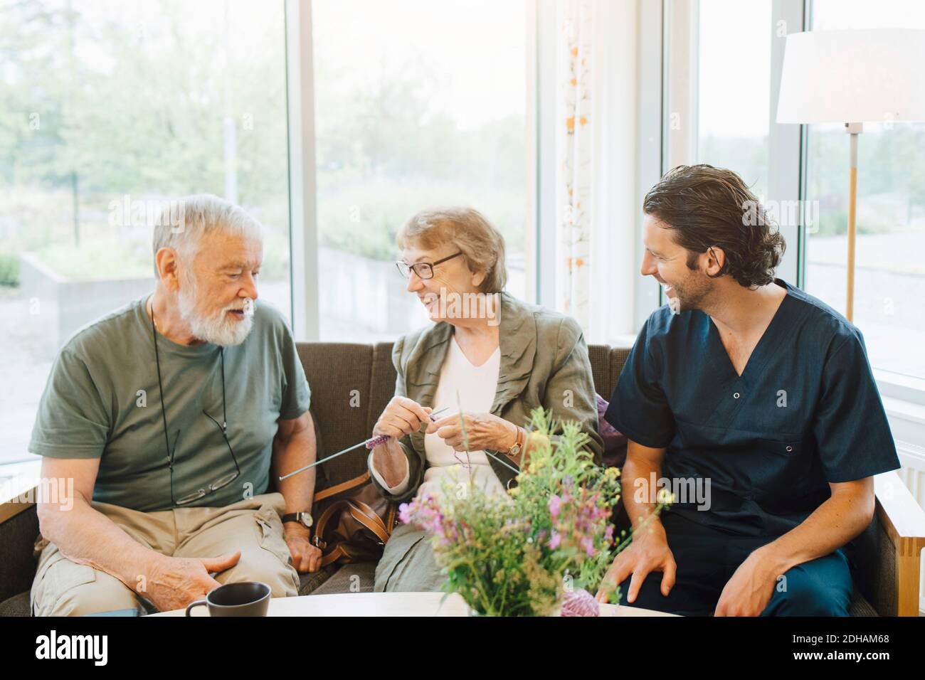 Smiling retired man and woman talking with male caregiver while sitting on sofa against window at nursing home Stock Photo