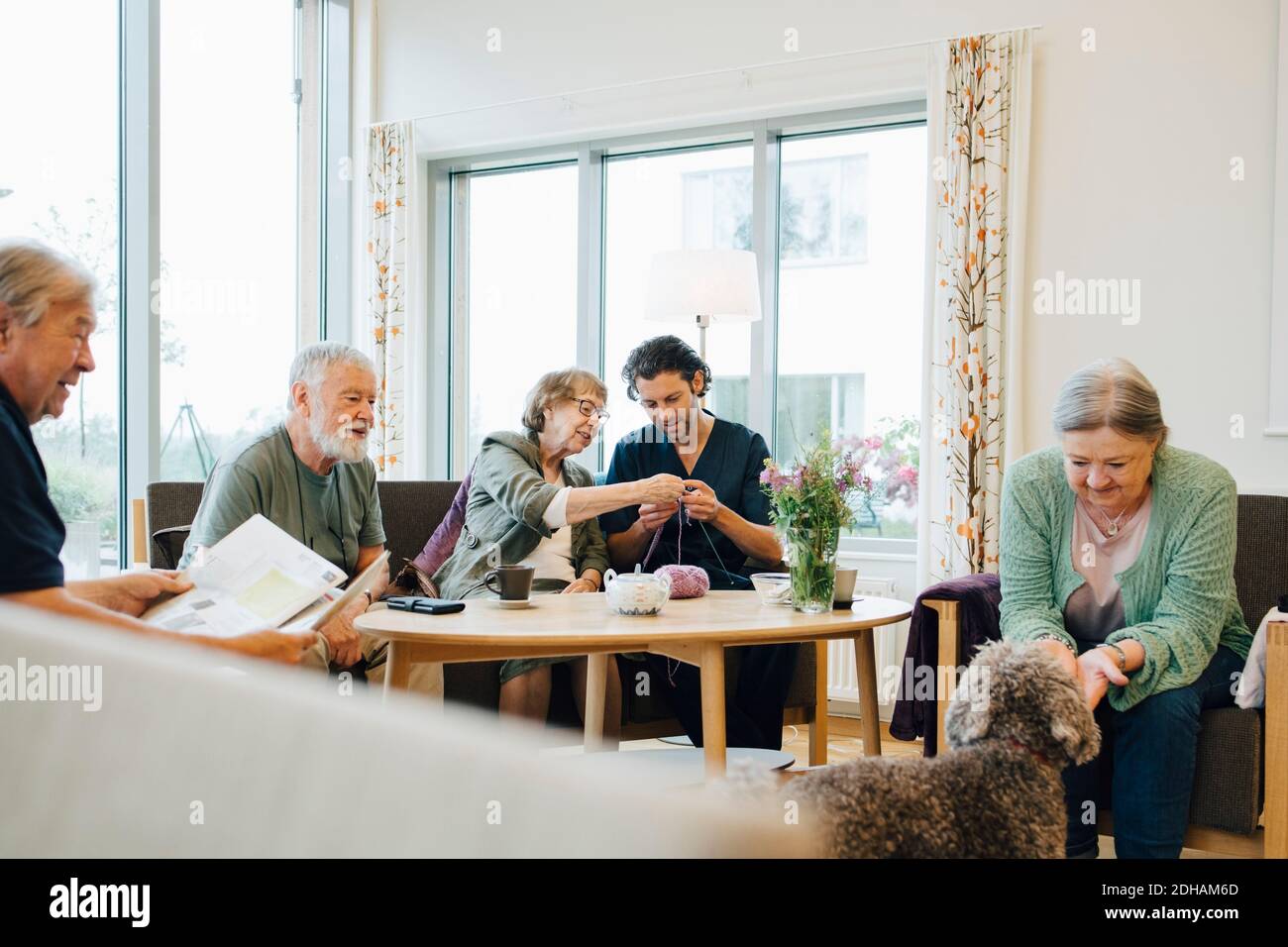 Retired senior woman teaching knitting to male nurse while sitting amidst friends at elderly care home Stock Photo
