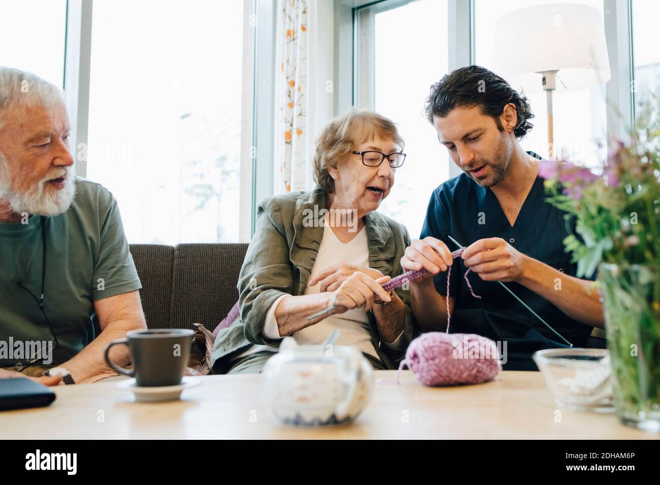 Retired senior woman teaching knitting to male nurse while sitting on sofa at elderly care home Stock Photo