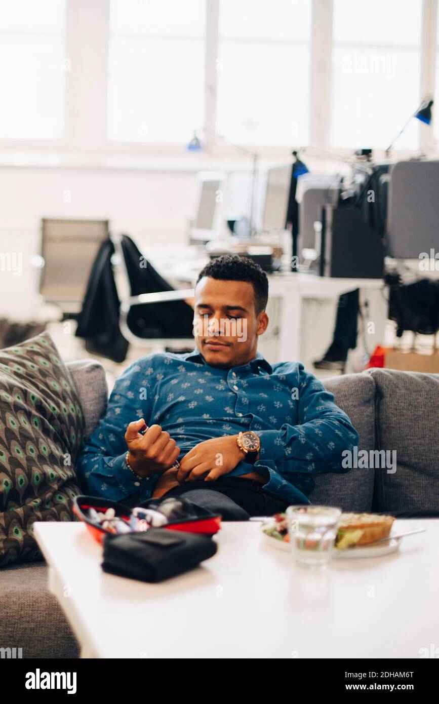 Businessman injecting insulin while sitting on sofa in office Stock Photo