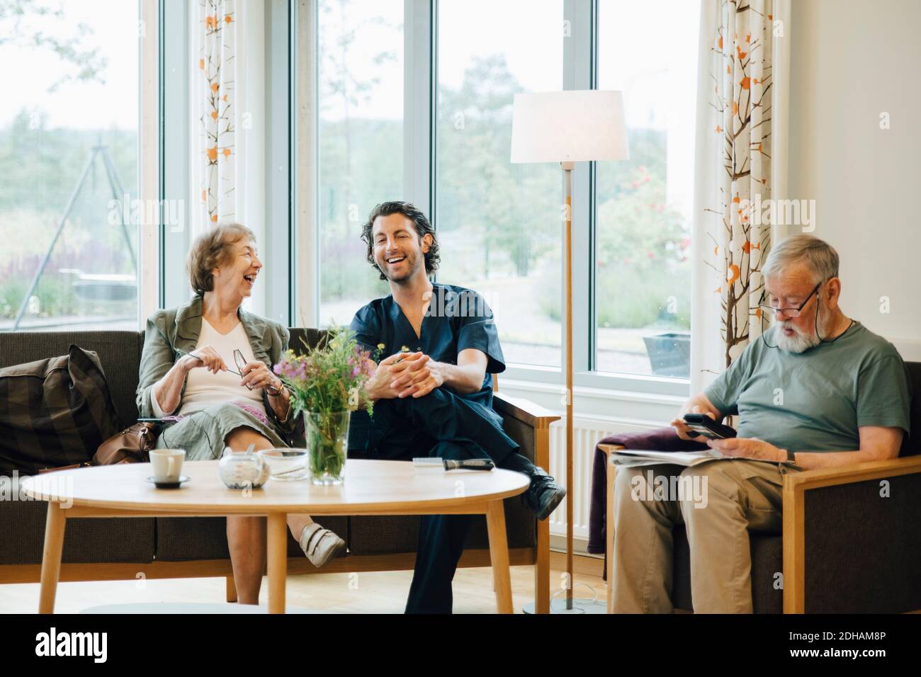 Cheerful senior woman talking with male nurse sitting on sofa against window at nursing home Stock Photo