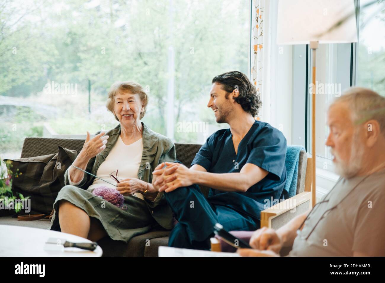 Smiling senior woman talking with male nurse sitting on sofa against window at nursing home Stock Photo