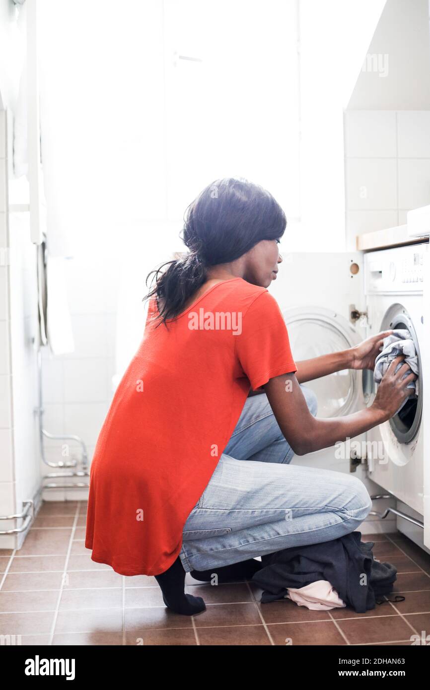 Full length side view of mid adult woman putting clothes into washing machine at home Stock Photo