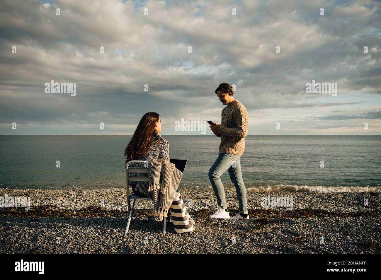 Man using mobile phone while woman sitting with laptop on chair at beach against sky Stock Photo
