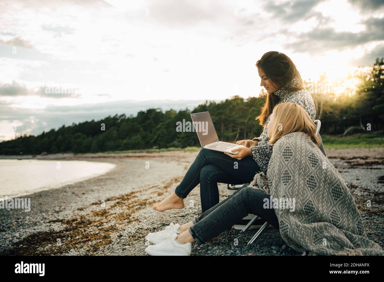Woman wrapped in blanket looking at friend using laptop on sea shore Stock Photo