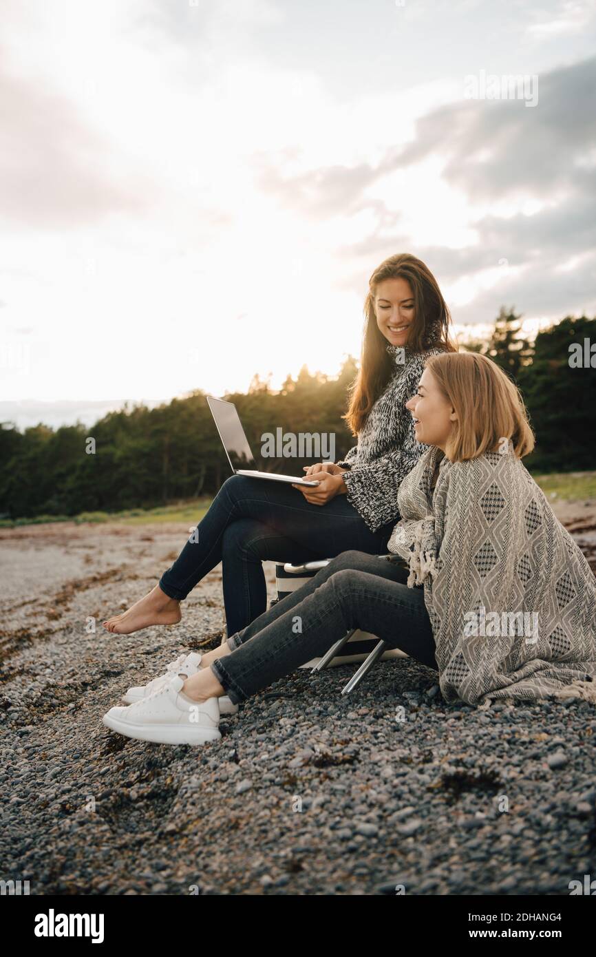 Smiling woman holding laptop while sitting with friend at beach during cold weather Stock Photo