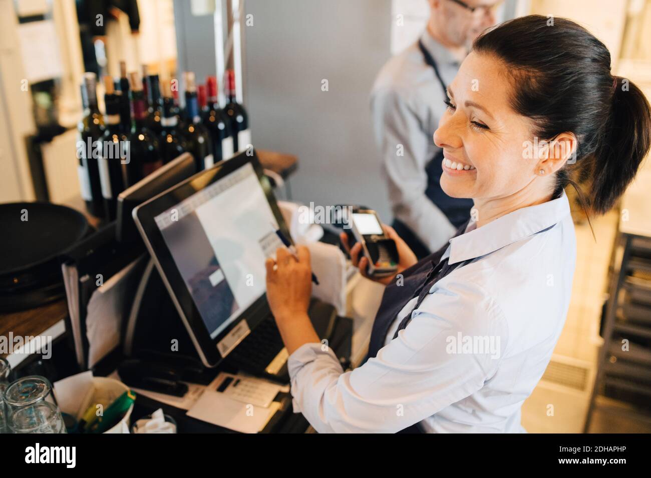 Smiling woman using computer while holding credit card reader in restaurant Stock Photo