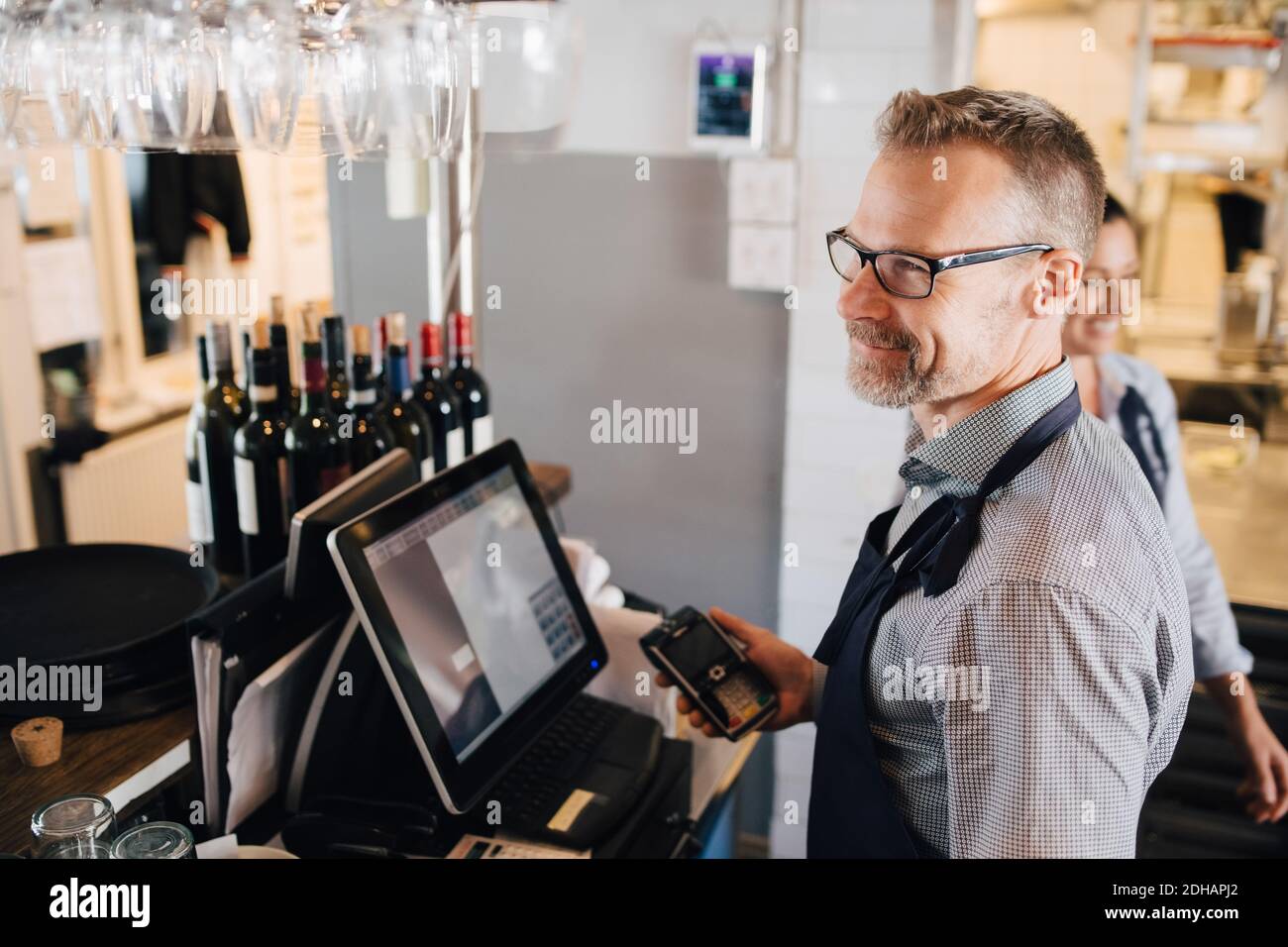 Smiling man using computer while holding credit card reader in restaurant Stock Photo