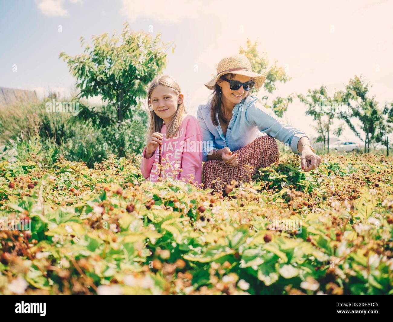 Smiling woman and daughter sitting amidst plants at farm against sky on sunny day Stock Photo