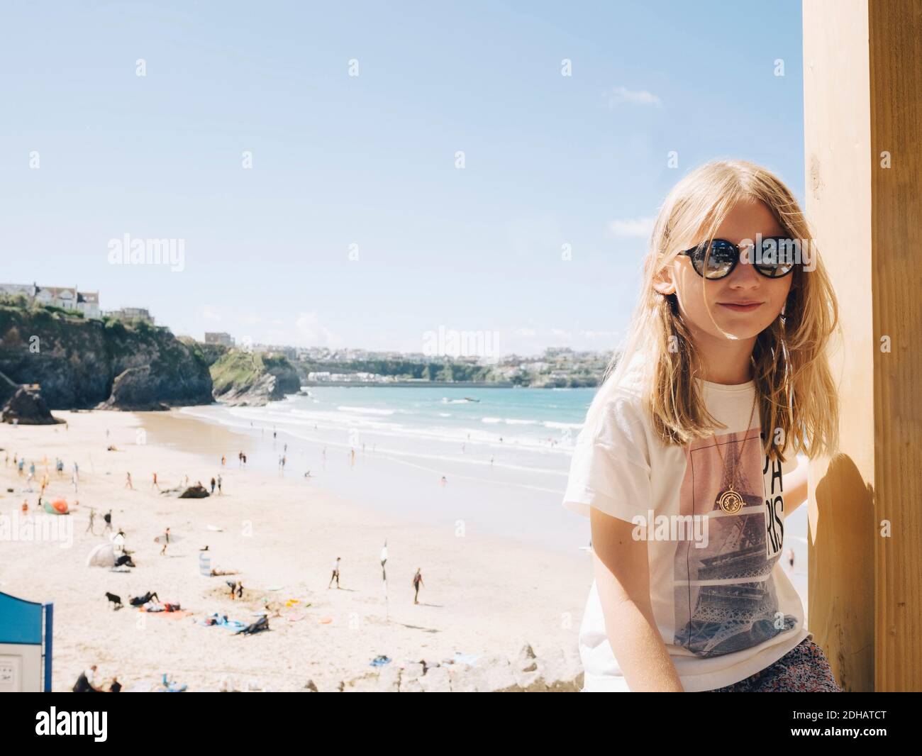 Blond girl sitting by wooden column at beach against sky on sunny day Stock Photo