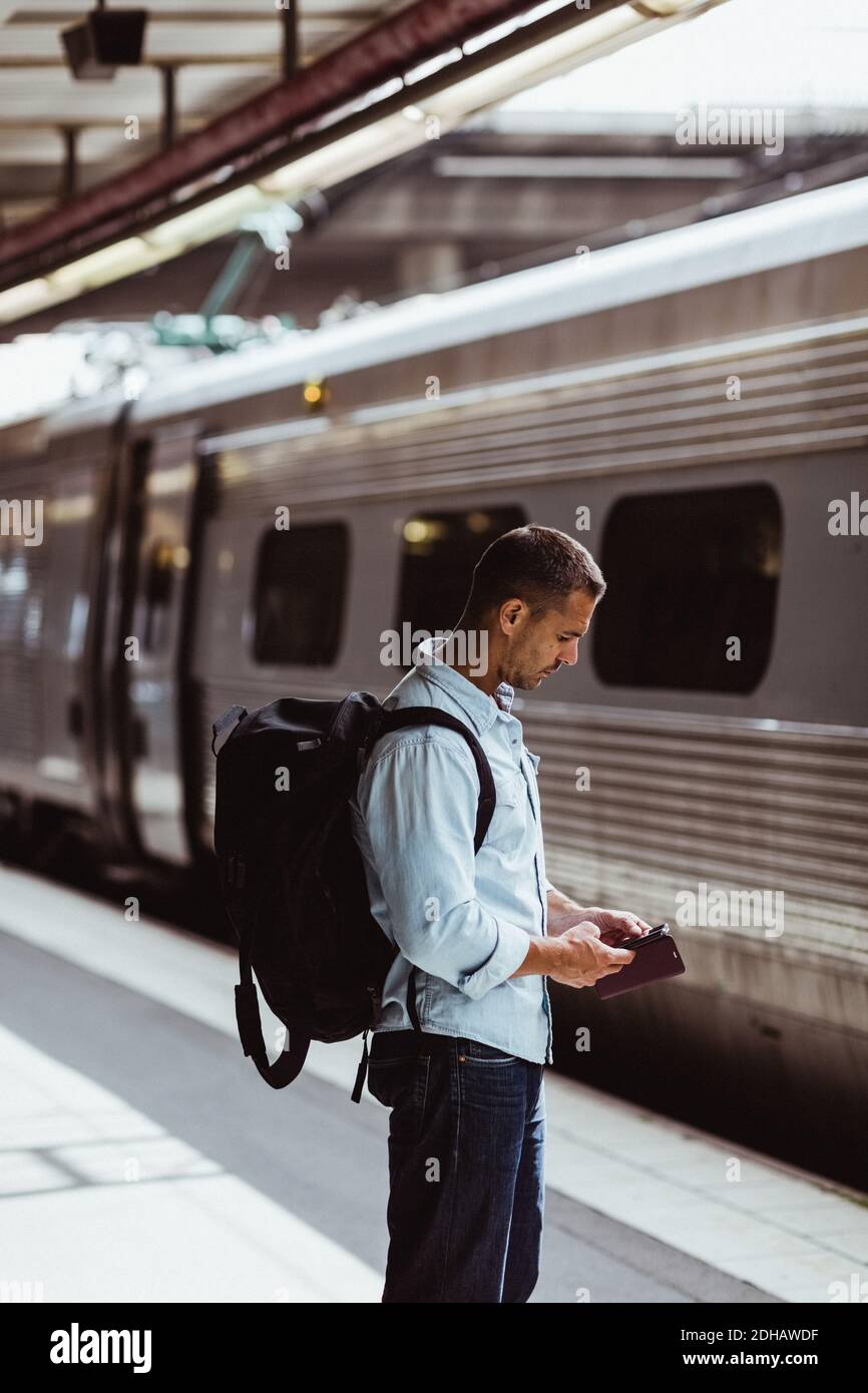 Mid adult man with backpack using mobile phone while standing by train on platform Stock Photo