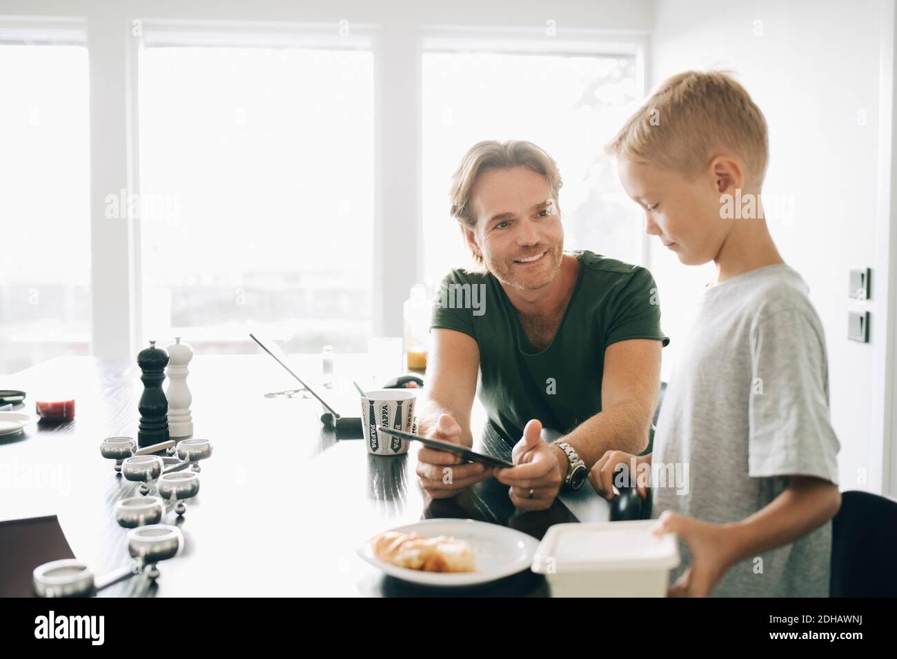 Smiling man showing digital tablet to boy at dining table against window Stock Photo