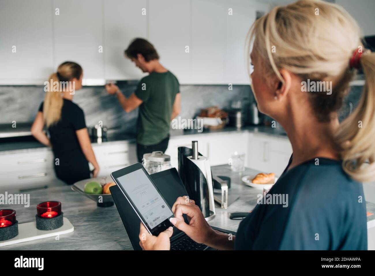 Mature woman with technologies looking at man and daughter standing in kitchen Stock Photo