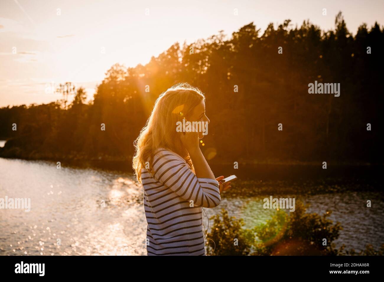 Side view of mid adult woman using mobile phone while standing against lake at sunset Stock Photo