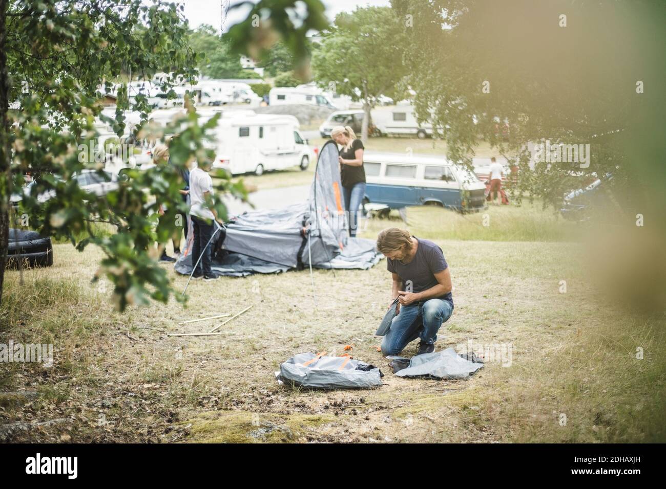 Father searching in bag while family pitching tent in background at campsite Stock Photo
