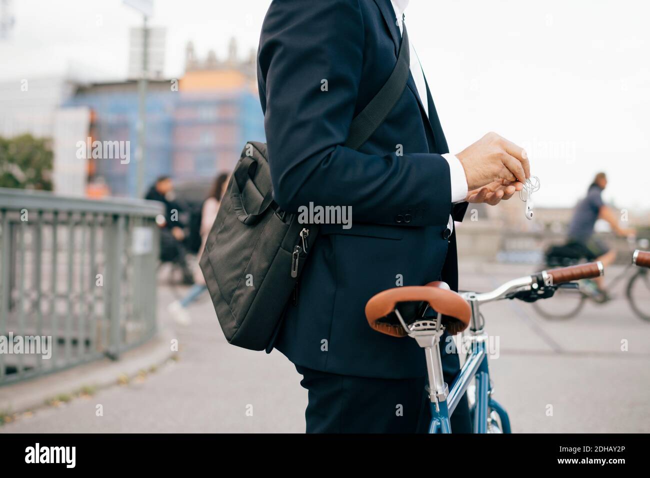 Midsection of businessman holding in-ear headphones while standing with bicycle on bridge in city Stock Photo