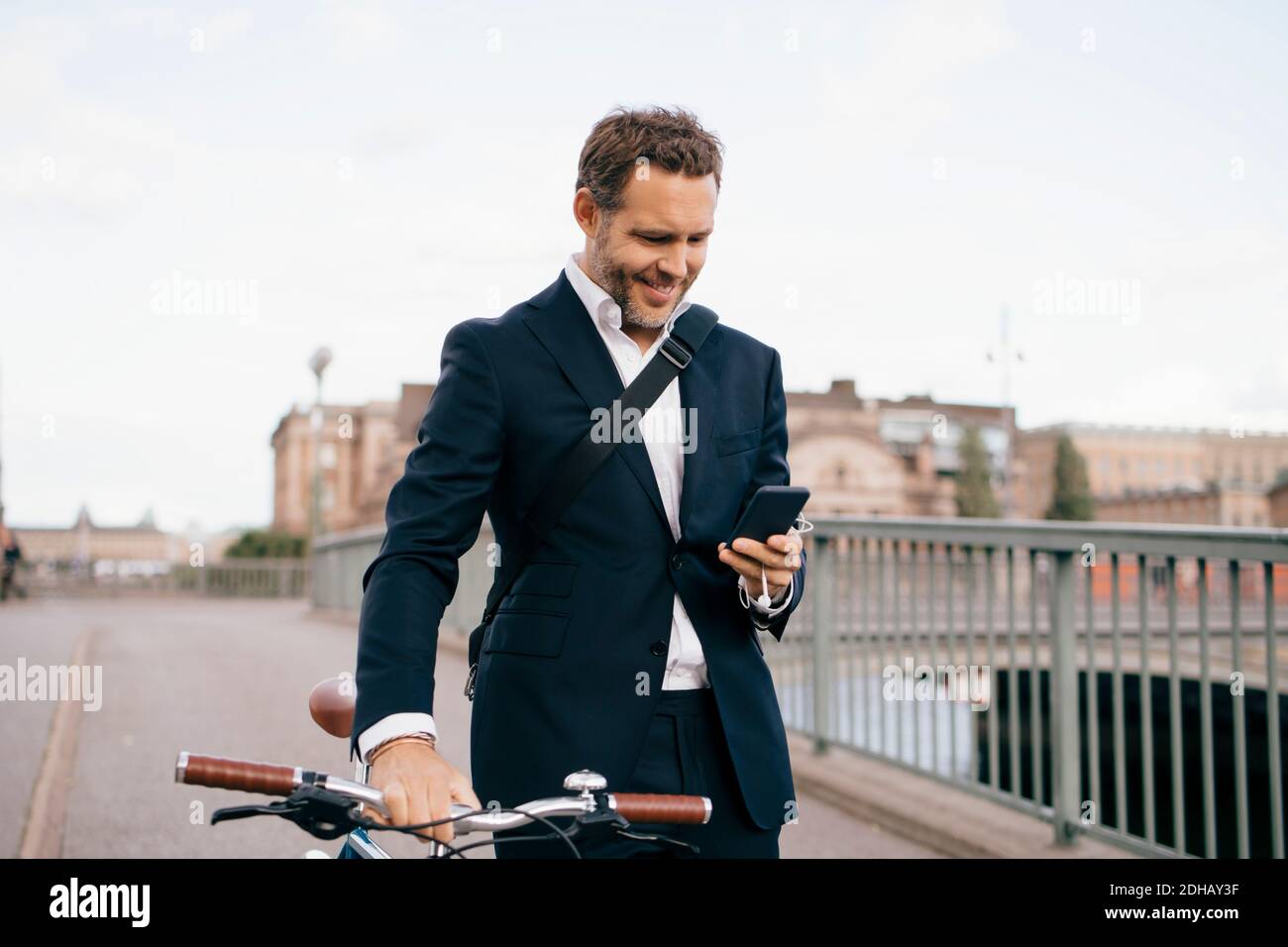 Smiling businessman using mobile phone while walking with bicycle on bridge in city Stock Photo
