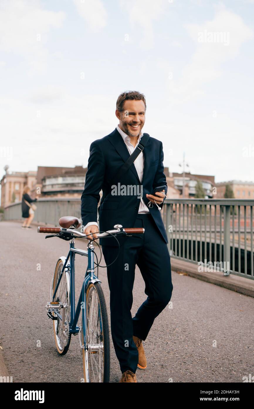 Smiling businessman holding smart phone while walking with bicycle on bridge in city Stock Photo