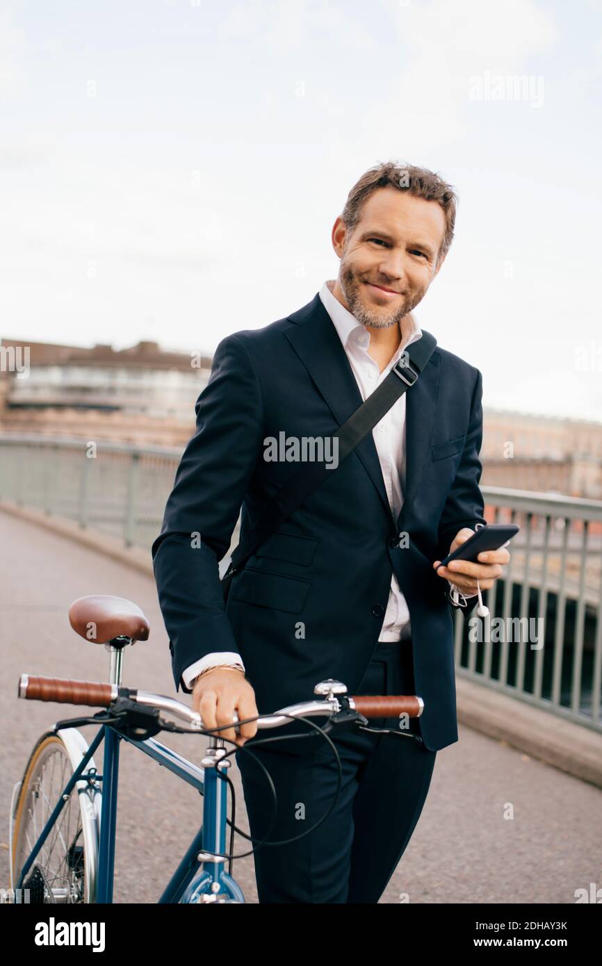 Portrait of confident businessman holding mobile phone while walking with bicycle on bridge in city Stock Photo
