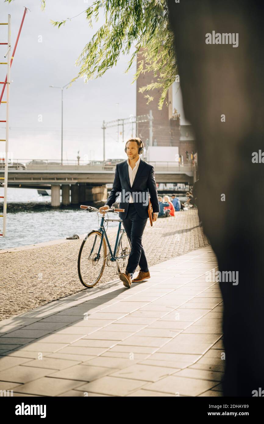 Businessman walking with bicycle on footpath in city during sunny day Stock Photo