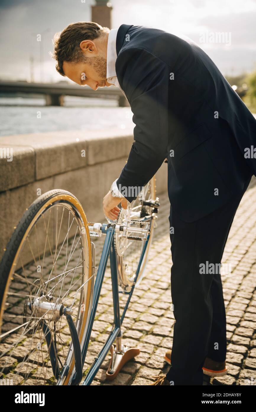 Side view of businessman repairing bicycle on footpath in city during sunny day Stock Photo