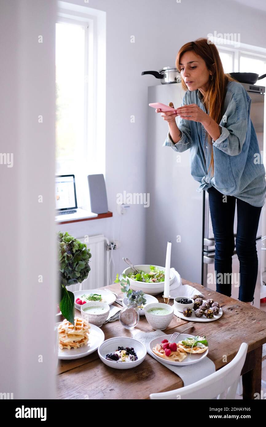 Blogger photographing food at table through mobile phone by window Stock Photo