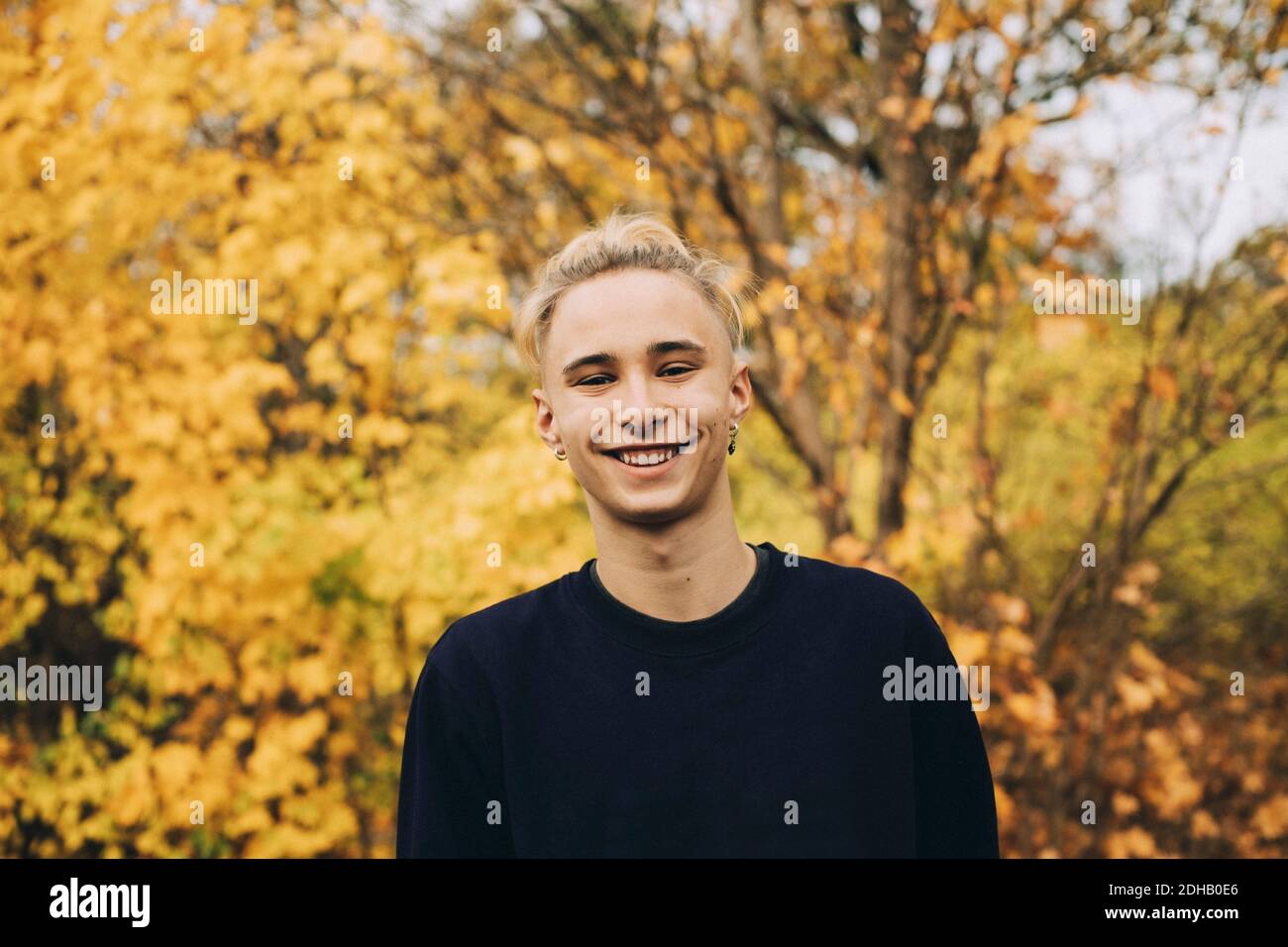 Portrait of smiling teenage boy with blond hair standing against maple trees Stock Photo
