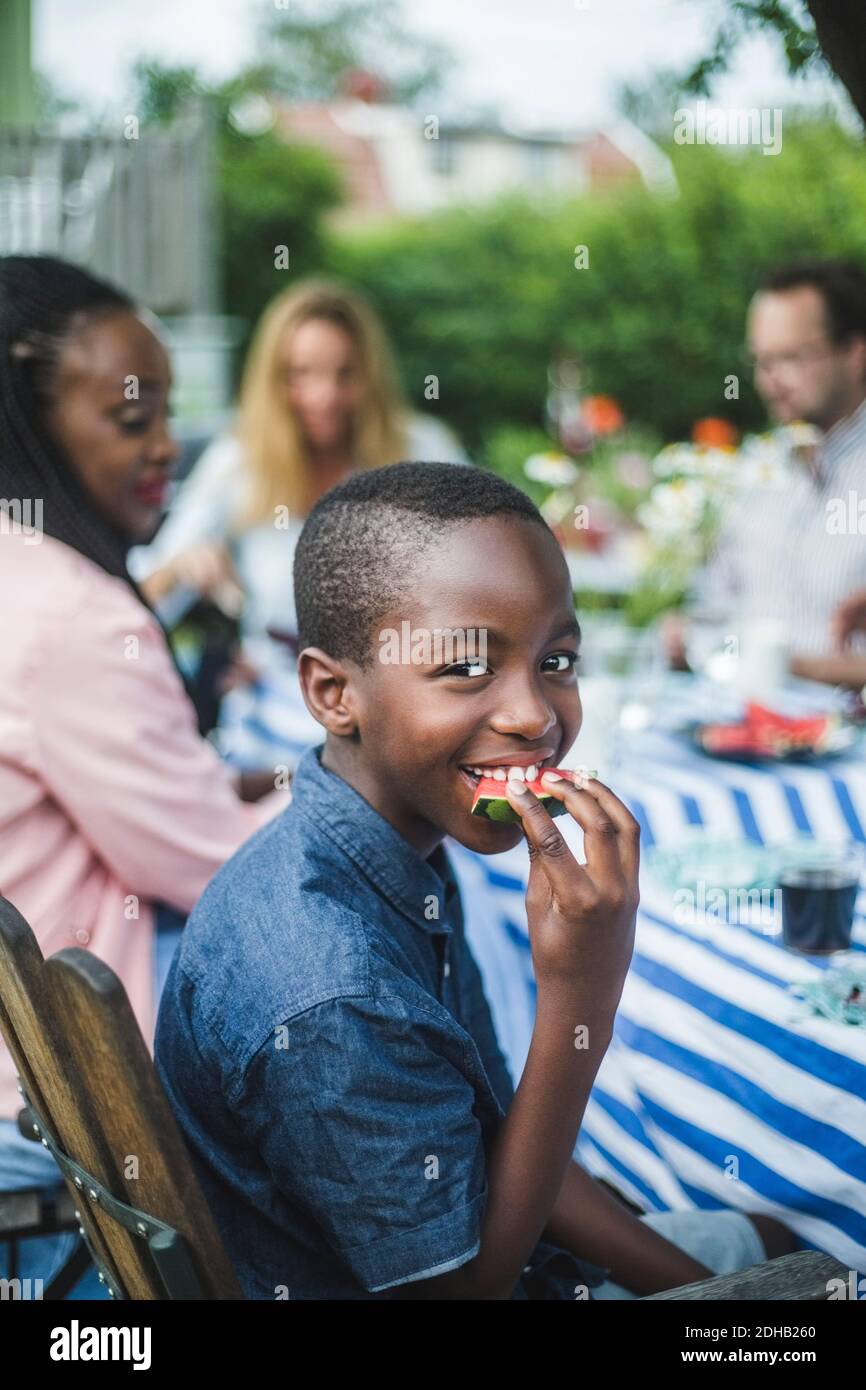 Smiling boy eating watermelon while sitting with family at table in garden Stock Photo