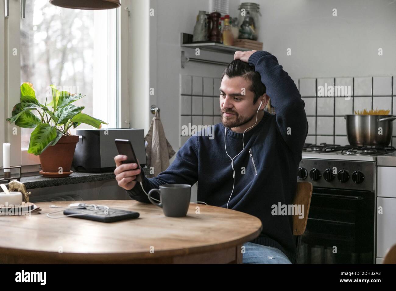 Man with headphones using mobile phone while sitting at table in kitchen Stock Photo