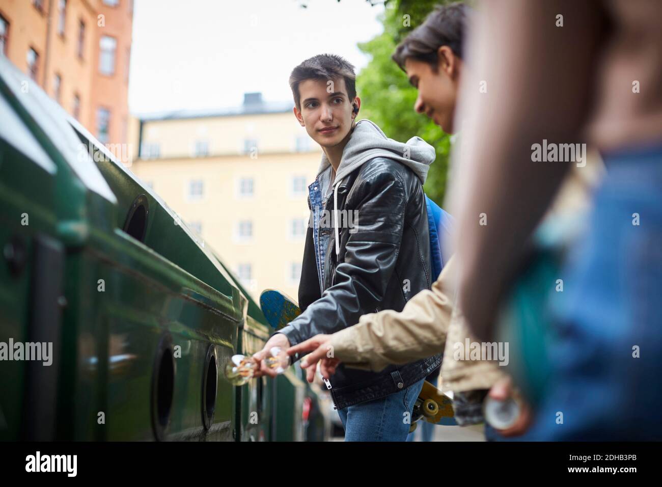 Teenage boy looking at friend while throwing waste in garbage bin at recycling station Stock Photo