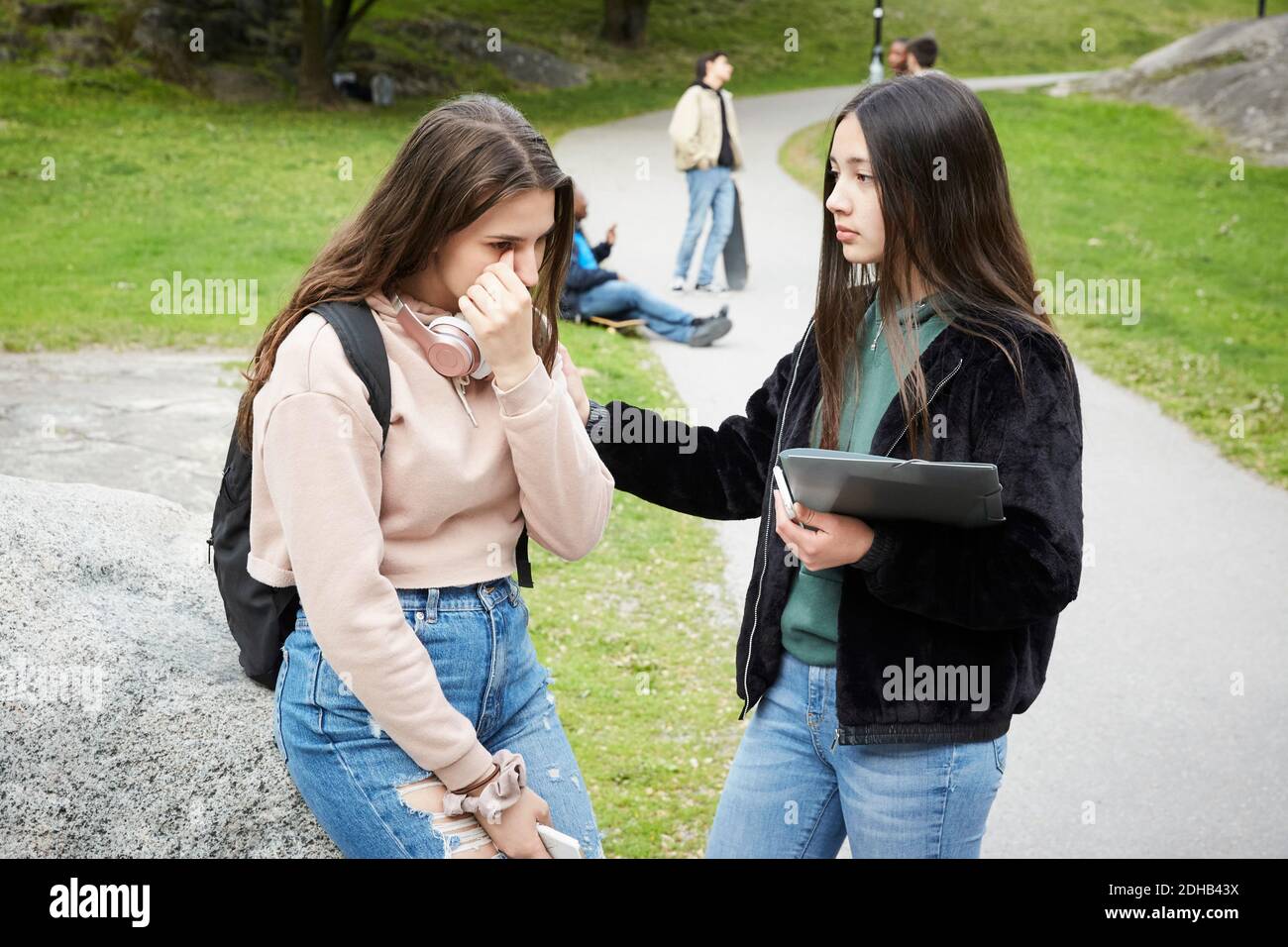 Teenage girl looking at female friend crying while sitting on rock at park Stock Photo