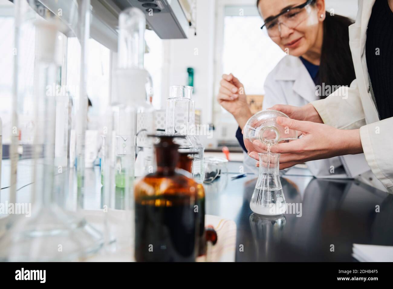 Midsection of young female student pouring solution in flask by teacher at chemistry laboratory Stock Photo